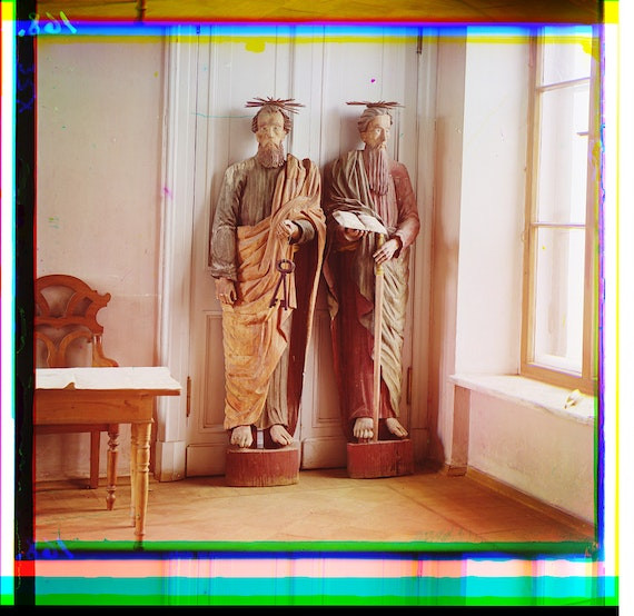 Photograph of two statues in front of a door. The photo is of orange-brown tint. The frame/fringes separate into color planes, like wet rainbows. Top is red--magenta-cyan-blue-green-yellow, bottom is more off, green-pink-cyan-green again.
