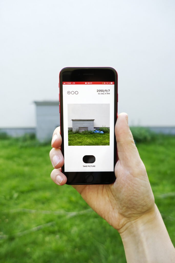 photo: hand holding a smartphone, which shows a camera-like app; the viewfinder window shows the scene behind the smarthpone: a tiny concrete box structure in front of a white wall and grass