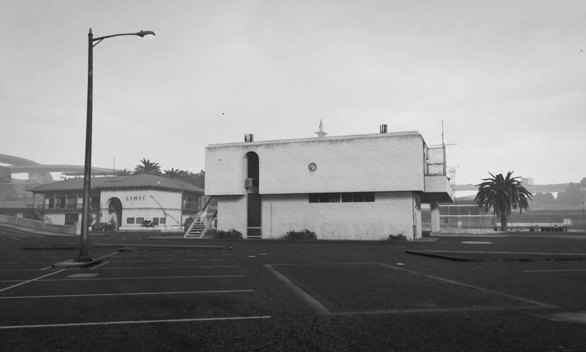 A black-and-white landscape "photograph" of a non-descript, oncrete-heavy building. The second floor makes it look slightly mushroom-shaped. In the front, big parking lot, a street lamp. In the background palm trees and a building that says LSMYC. If you look close enough you realize that this is not a photograph, but a screengrab.