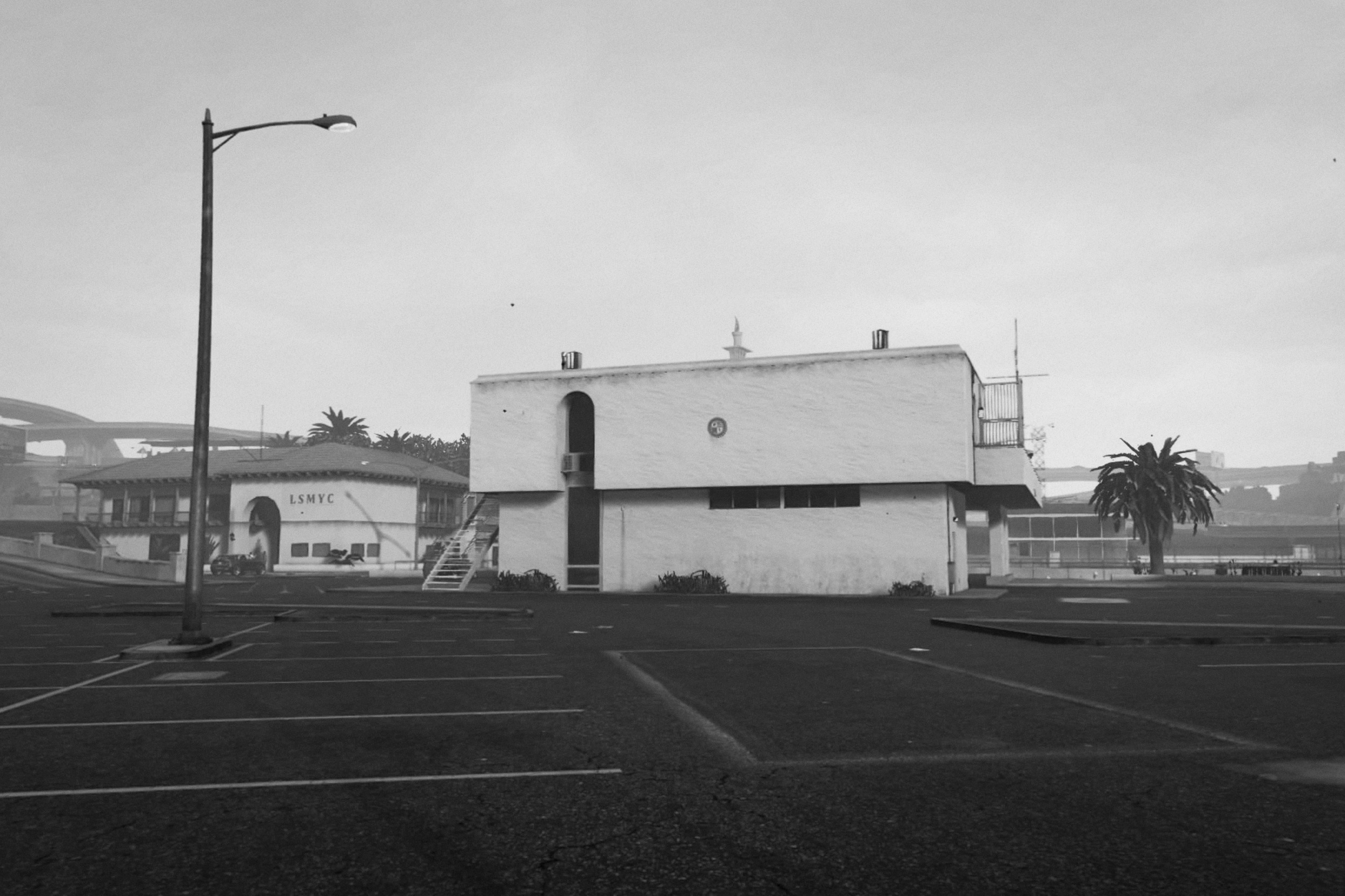 A black-and-white landscape "photograph" of a non-descript, oncrete-heavy building. The second floor makes it look slightly mushroom-shaped. In the front, big parking lot, a street lamp. In the background palm trees and a building that says LSMYC. If you look close enough you realize that this is not a photograph, but a screengrab. 