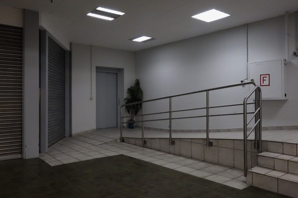 Photograph: a dark, neon-lit backroom, maybe in a mall. There are shop window blinds, an elevator door, a fire extinguisher box, and a sad plant in the corner. The floor is dark carpet and grey dirty tiles. A steel guardrail strongly guards a ramp that leads out of the picture.