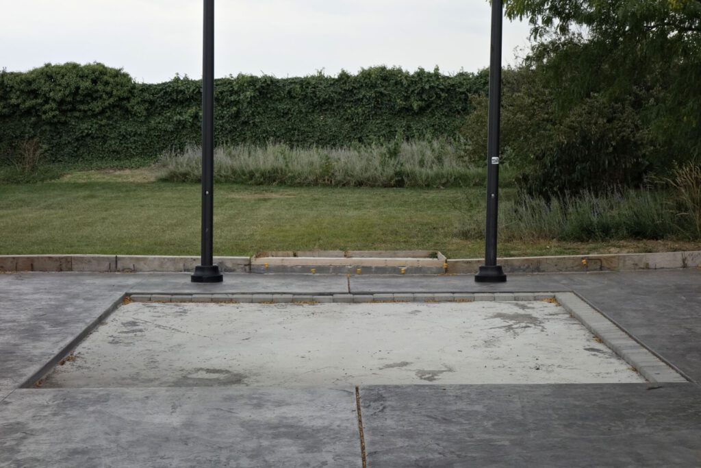 Photograph: a slightly surreal, very symmetrical scene: two lamp posts between a grass and hege background, and a rectangular hole in the sidewalk which appears sandy. The sky is almost white.
