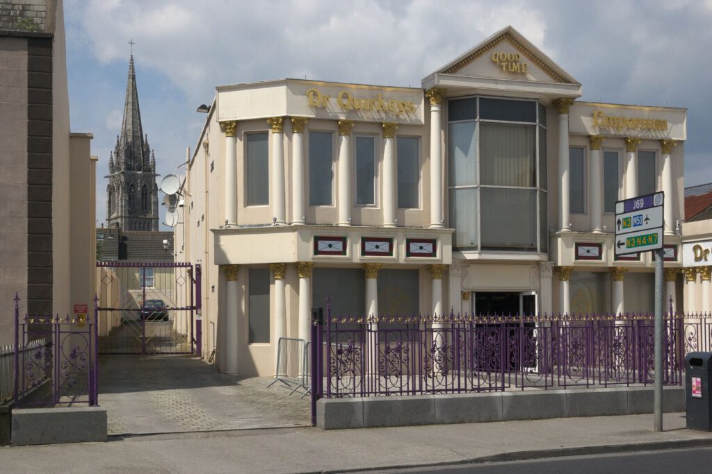Photograph: a strange building in a residential area. Two storey, but lots of columns, a purple fence and golden letters. They are hard to read but say "Dr Quirkeys emporium - Good Time". At the end of the back alley you can see the tower of a cathedral.