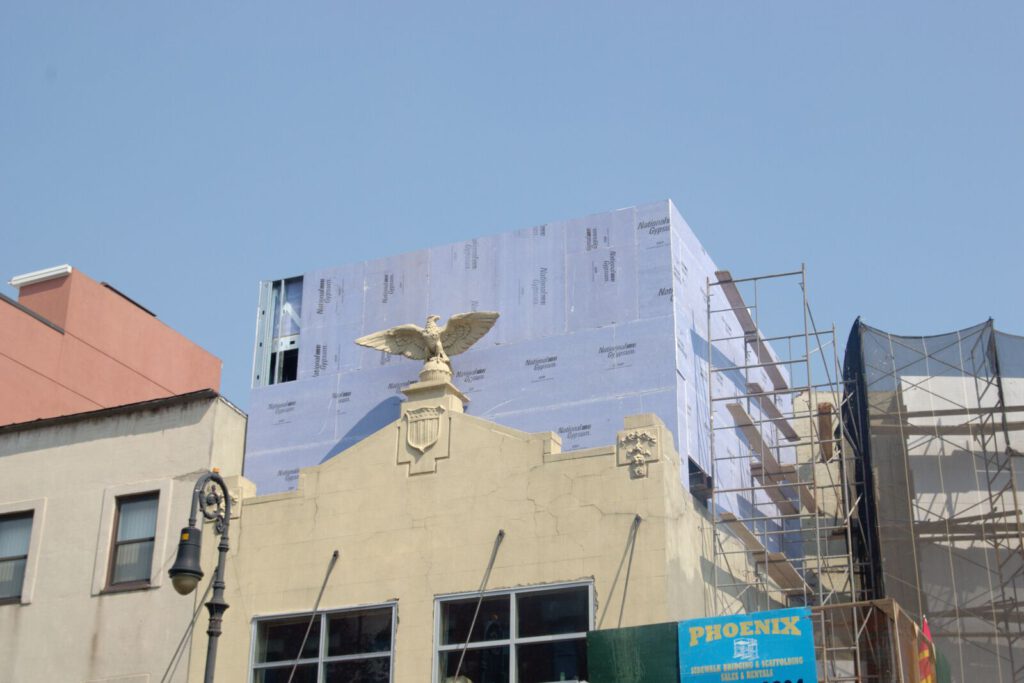 Photograph: Construction on top of a small building: the roof is covered by purplish panels which makes it blend almost into the pale blue sky. In front of the purplish backdrop, a majestic eagle sculpture. In the corner, a sign advertises a company named "Phoenix". The image has a pastel palette.