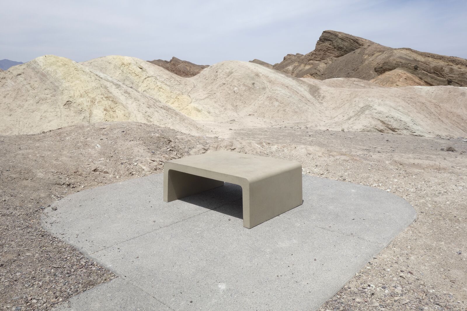 Photograph: a minimalist concrete bench on a rounded patch of concrete, surrounded by a rocky, mountain vista. Everything is very beige, the sky is pale blue.