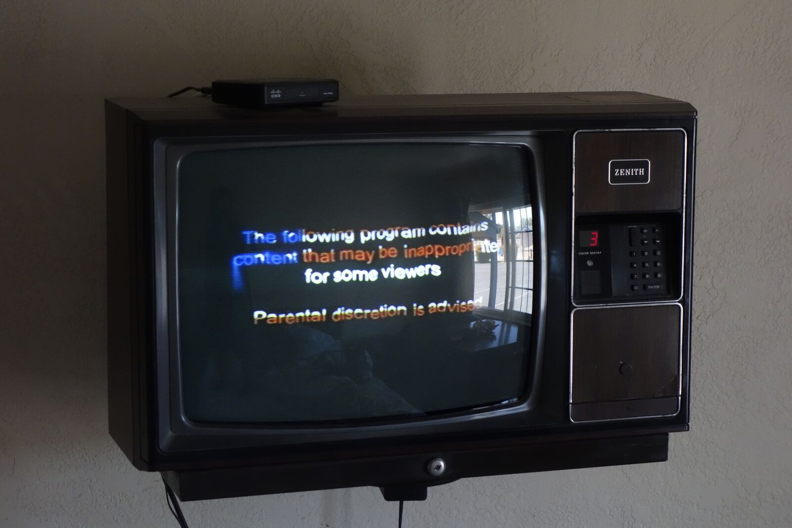 Photograph: an 80s "Zenith" TV mounted on the wall. The screen shows a distorted text, a parental discretion warning; the text is colored as the US flag.