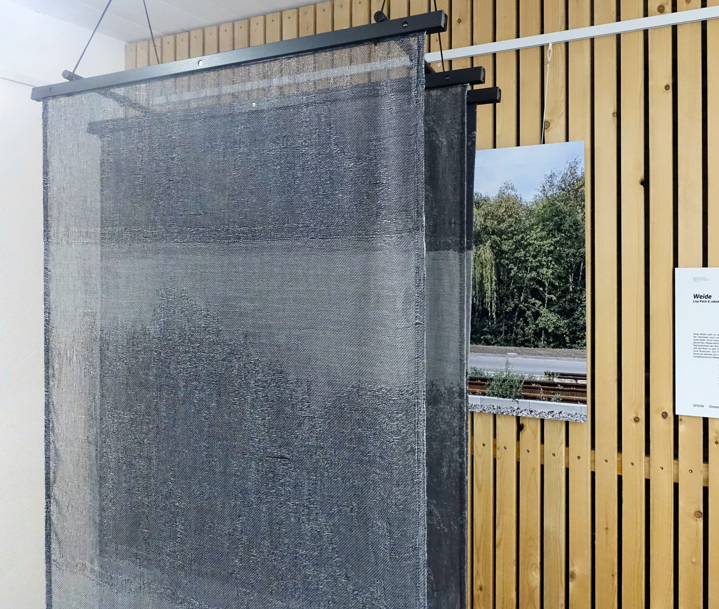 Three layers of black and white woven textiles, hanging vertically, parallely, in front of a glossy photograph on the wall. The wall looks like a sauna. On the photograph, we can see a willow tree, some sky, and road in front of it.