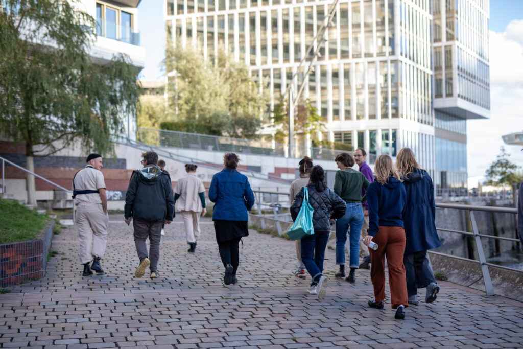 photo: a tour guide in khaki overalls leading a group of participants (we see their backs) towards a skyscraper or office building