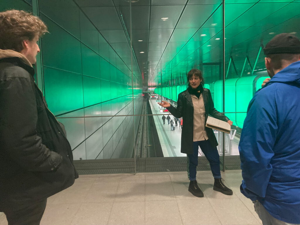 photo: a tour guide gesturing to the place we're at: a magically green lit subway station. Two people listen to her.
